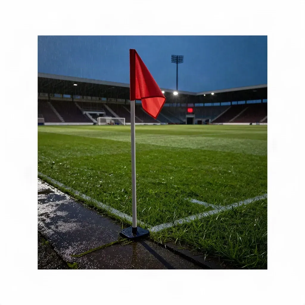 Campo da calcio bagnato, bandierina rossa, tribune illuminate dopo la pioggia al tramonto.